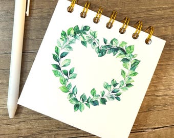 May include: A square spiral-bound notebook with a white cover featuring a watercolor illustration of a heart-shaped wreath made of green leaves, accompanied by a plain white retractable ballpoint pen with a pocket clip.