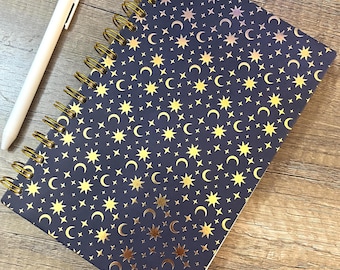 May include: A dark blue spiral-bound notebook featuring a repeating pattern of gold-colored stars and crescent moons, accompanied by a plain white retractable ballpoint pen positioned to the left of the notebook on a wood-grain surface.