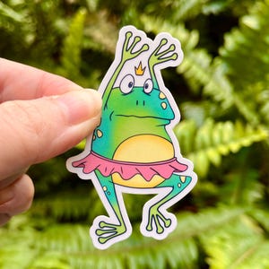 May include: A whimsical sticker featuring a cartoon frog in a pink tutu and a tiny gold crown. The frog is green with a yellow belly and is posed with its arms raised. The sticker is held against a backdrop of green foliage.
