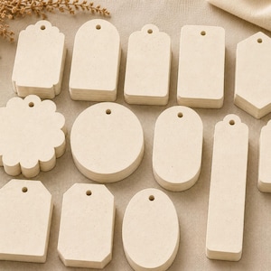 May include: A collection of twelve stacks of off-white, speckled paper tags in various geometric shapes, including circles, rectangles, ovals, and scalloped edges. Each tag features a small, centered hole at the top for stringing or attachment.