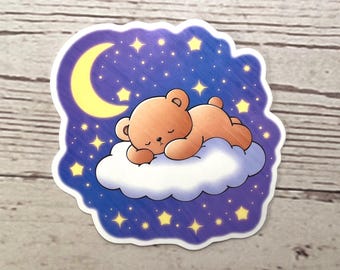 May include: A sticker featuring a cartoon bear sleeping on a cloud under a crescent moon and stars. The bear is light brown, the cloud is white, and the background is a gradient of blue and purple. Yellow stars and a yellow moon are also present.