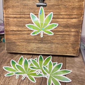 May include: A set of five green and white holographic marijuana leaf stickers. The stickers are arranged in a pile on a wooden surface.