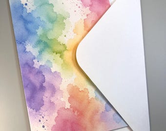 May include: A rectangular greeting card featuring a vibrant watercolor wash in a rainbow spectrum of pink, orange, yellow, green, blue, and purple hues, paired with a plain white envelope tucked behind the card for a clean presentation.