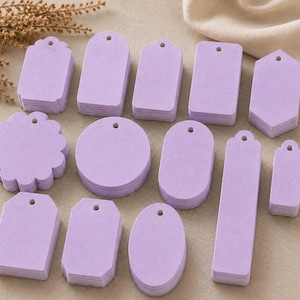 May include: A collection of light purple paper tags in various shapes, including circles, rectangles, ovals, and scalloped edges. Each tag features a small pre-punched hole at the top for stringing, arranged in multiple stacks on a flat surface.