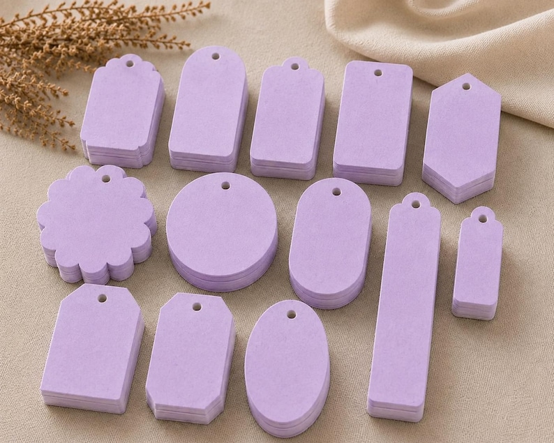 May include: A collection of light purple paper tags in various shapes, including circles, rectangles, ovals, and scalloped edges. Each tag features a small pre-punched hole at the top for stringing, arranged in multiple stacks on a flat surface.
