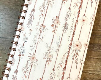 May include: A spiral-bound notebook with a white cover featuring a repeating pattern of vertical ropes in a reddish-brown hue, accented with delicate illustrations of soft pink roses, small daisies, and muted green foliage throughout the design.