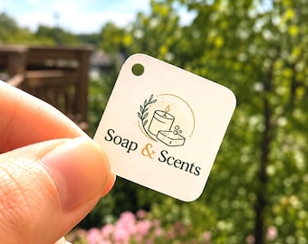 May include: A square white tag with rounded corners and a small hole at the top. The tag features a logo with a line-art candle and a bar of soap, surrounded by a leafy branch and a circle. The text Soap & Scents is printed in black and gold.