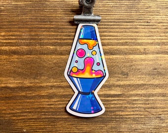 May include: A sticker featuring a blue lava lamp design with pink, orange, and yellow liquid. The sticker has a white border and is held by a metal clip. The background is a wooden surface.