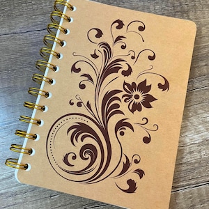 May include: A spiral-bound notebook with a tan cover featuring a dark brown floral and scrollwork design. The notebook has a gold-colored metal wire binding along the left side and rounded corners on the rectangular cover.
