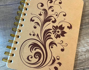 May include: A spiral-bound notebook with a tan cover featuring a dark brown floral and scrollwork design. The notebook has a gold-colored metal wire binding along the left side and rounded corners on the rectangular cover.