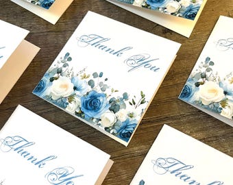 May include: A collection of square greeting cards featuring the text Thank You in elegant blue script. Each card is decorated with a watercolor illustration of white and blue roses, along with green eucalyptus leaves arranged along the bottom edge.