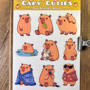 May include: A mini sticker sheet featuring cartoon capybaras in various poses. The stickers include capybaras with a leaf, drinking boba, wearing sunglasses, and wrapped in a blue blanket. The sheet has the text "CAPY CUTIES" at the top.