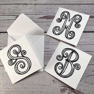 May include: White square cards with black swirly letter designs, including the letters M, B, and S, are displayed with two white envelopes on a light wood grain surface. The cards are blank and ready for personalization.