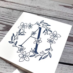 May include: A square white greeting card featuring a dark blue printed design of the letter A centered within a circular wreath of line-drawn flowers, leaves, and small berries. The card is folded and displays a clean, minimalist graphic.