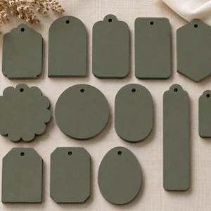May include: A collection of twelve olive green tags in various geometric shapes, including circles, rectangles, ovals, and scalloped edges. Each tag features a single small circular hole at the top, designed for stringing or attachment to various items.