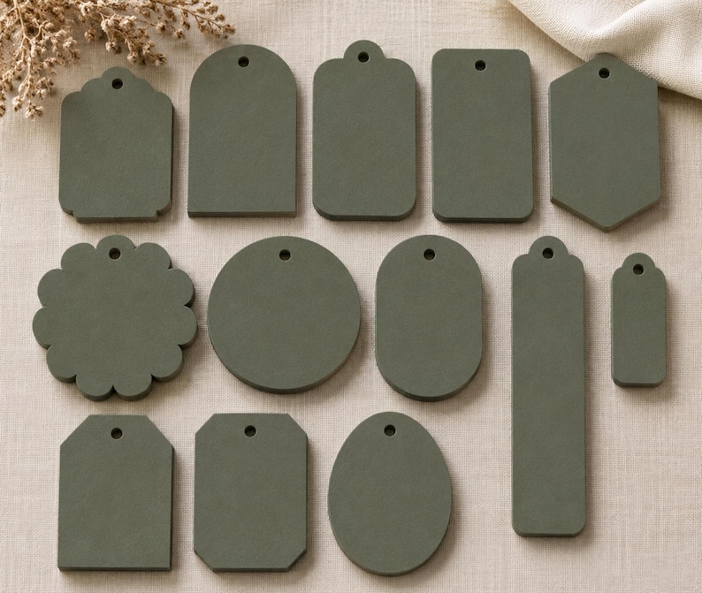 May include: A collection of twelve olive green tags in various geometric shapes, including circles, rectangles, ovals, and scalloped edges. Each tag features a single small circular hole at the top, designed for stringing or attachment to various items.