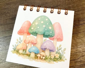May include: A square spiral-bound notepad featuring a watercolor illustration of various mushrooms in pastel shades of green, pink, purple, and blue, surrounded by small green leaves and tiny yellow flowers on a white background.