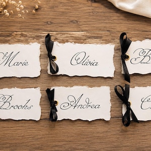 May include: Six rectangular white paper tags with deckled edges, each featuring a black metal eyelet and a tied black satin ribbon. The names Marie, Olivia, Benjamin, Brooks, Andrea, and Chelsea are printed in a black cursive script font.
