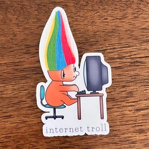 May include: A sticker of a cartoon troll with rainbow hair sitting at a computer desk. The troll is wearing an orange shirt and is typing on a keyboard. The sticker says "internet troll" at the bottom.