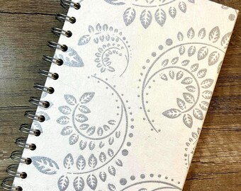 May include: A spiral-bound notebook with a cream-colored cover featuring a repeating pattern of silver-toned, curved leafy vine motifs. The notebook is secured with a black metal wire coil along the left side, providing a functional binding.