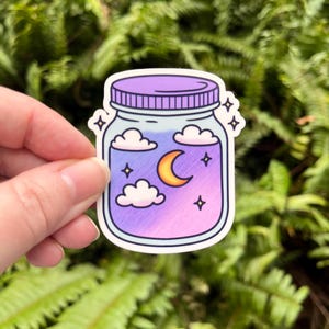 May include: A sticker featuring a jar with a purple lid and a night sky scene inside. The jar contains a crescent moon, clouds, and stars, with a gradient of purple and pink hues. The sticker has a white border and is held against a backdrop of green foliage.