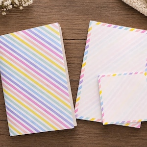 May include: A stationery set consisting of a notebook, a rectangular sheet of paper, and a square sticky note, all featuring a pastel diagonal striped pattern in shades of pink, yellow, blue, and purple on a white background.