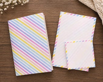 May include: A stationery set consisting of a notebook, a rectangular sheet of paper, and a square sticky note, all featuring a pastel diagonal striped pattern in shades of pink, yellow, blue, and purple on a white background.