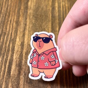 May include: A sticker featuring a cartoon capybara wearing sunglasses and a red Hawaiian shirt with white flower patterns. The capybara is standing on a white background.