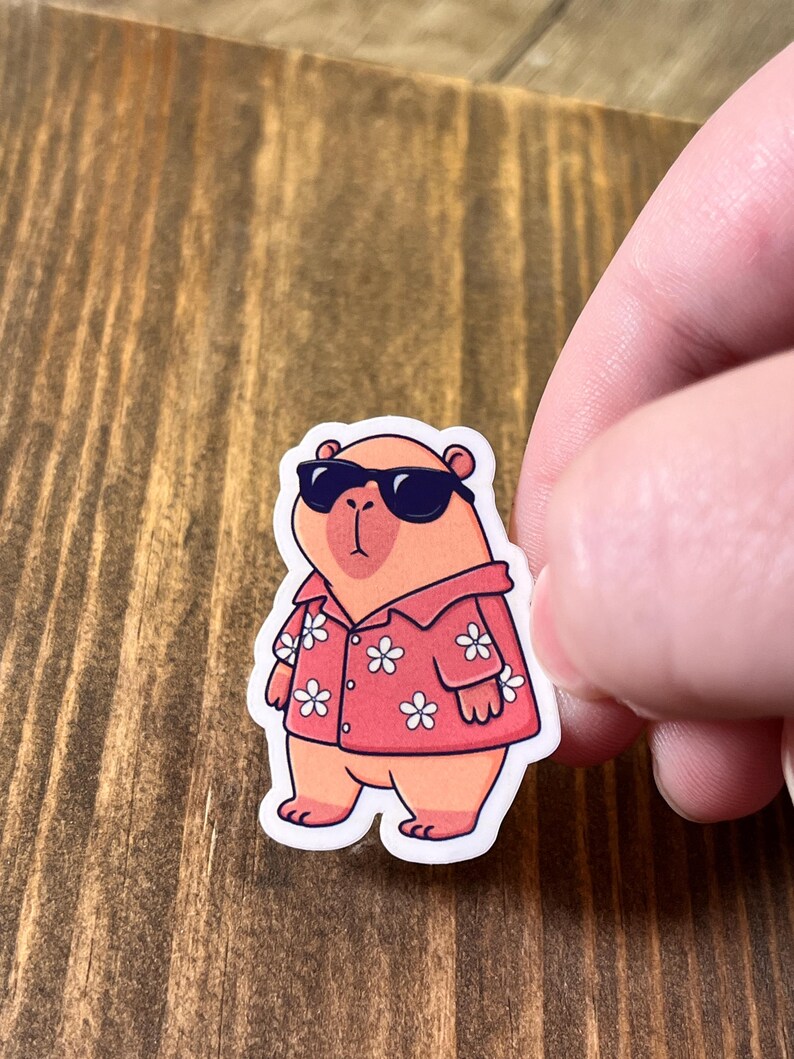 May include: A sticker featuring a cartoon capybara wearing sunglasses and a red Hawaiian shirt with white flower patterns. The capybara is standing on a white background.
