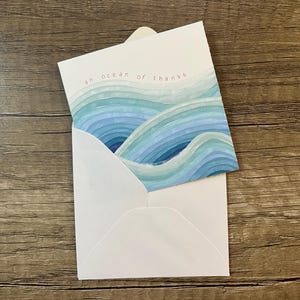 May include: A white greeting card tucked into a matching white envelope. The card features a watercolor illustration of blue and teal ocean waves with the text "an ocean of thanks" printed in small, simple, dark letters at the top center.