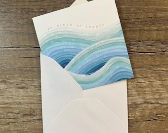 May include: A white greeting card tucked into a matching white envelope. The card features a watercolor illustration of blue and teal ocean waves with the text "an ocean of thanks" printed in small, simple, dark letters at the top center.