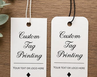 May include: Two rectangular white paper tags with rounded corners, each featuring the black printed text Custom Tag Printing and YOUR TEXT OR LOGO HERE with a small diamond icon. One tag has a white string and the other has a black string.