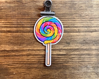 May include: A colorful lollipop sticker with a rainbow swirl design and a light brown stick. The sticker is held by a metal clip against a wooden background. The lollipop features stars.