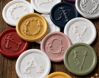 May include: A collection of circular wax seals in various colors including white, gold, red, navy blue, dusty pink, olive green, purple, and yellow. Each seal features an embossed letter or ornate floral frame design on the surface.