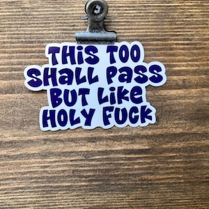 May include: A white sticker with the text "This too shall pass BUT Like HOLY FUCK" in blue, attached to a wooden surface with a metal clip.