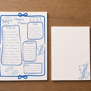May include: A white planner notepad titled Hanna's Planner with blue ink illustrations of cats, featuring sections for To Do List, Goals, Reminders, and Notes, alongside a matching blank notepad with a single cat illustration holding a pencil.