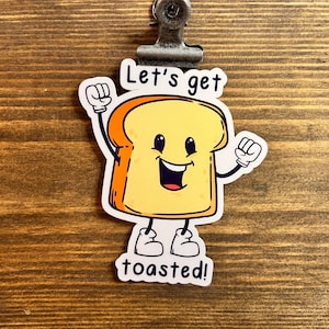 May include: A sticker featuring a cartoon slice of toast with a smiling face, arms raised, and feet. The text "Let's get toasted!" is above and below the toast illustration. The sticker is attached to a wooden surface.