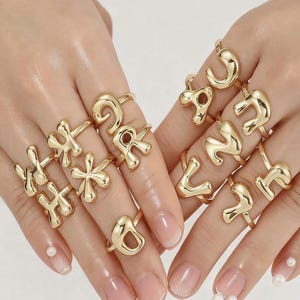 May include: A collection of gold-colored alphabet rings displayed on two hands. Each ring features a different letter of the alphabet in a bold, rounded font. The rings are shiny and reflect light, creating a glamorous look. The nails are painted with a light pink color.