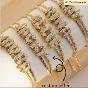 May include: Gold-toned bracelets with custom letter designs. Each bracelet features a series of sparkling, clear stones set in the shape of a letter. The bracelets are displayed on a light-colored surface, with the words "custom letters" at the bottom.