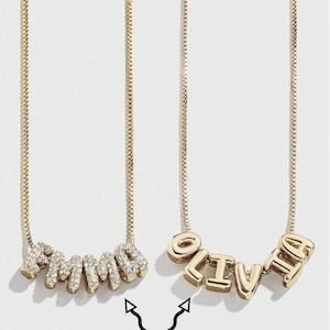 May include: Two gold-colored necklaces with custom text pendants. One necklace spells "emma" in a bubbly font with clear crystals. The other spells "olivia" in a block font. The text "Custom Text" is below the necklaces.