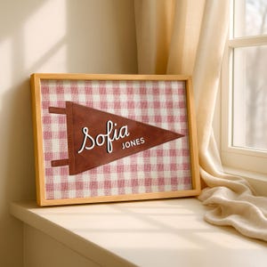 Może przedstawiać: Oprawiony proporczyk z napisem "Sofia Jones" białym skryptem na brązowym trójkącie. Tło to czerwono-biały wzór w kratę vichy. Rama wykonana jest z jasnego drewna.