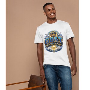 Op de afbeelding: Wit T-shirt met de tekst "GOAT God Over All Things" in blauw en goud. Het ontwerp bevat een kroon, duiven en een kruis. Het T-shirt wordt gedragen met een blauwe jeans.