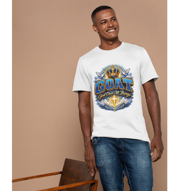 Op de afbeelding: Wit T-shirt met de tekst "GOAT God Over All Things" in blauw en goud. Het ontwerp bevat een kroon, duiven en een kruis. Het T-shirt wordt gedragen met een blauwe jeans.