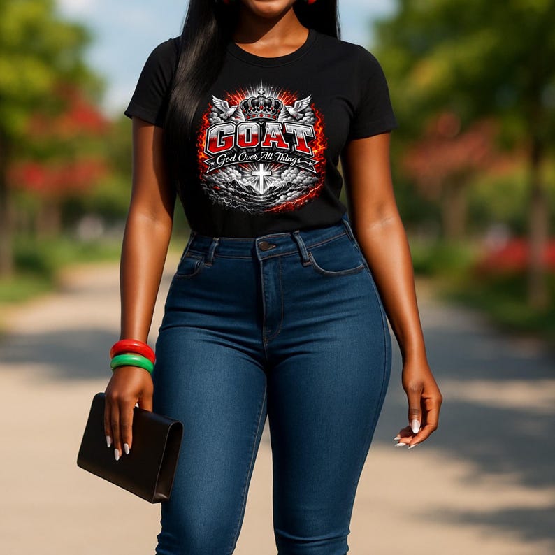 Op de afbeelding: Zwart T-shirt met ronde hals en de tekst "GOAT God Over All Things" in wit, rood en zilver. Het shirt wordt gecombineerd met een blauwe jeans. Een zwarte clutch en armbanden zijn ook zichtbaar.