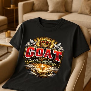 Op de afbeelding: Zwart t-shirt met het woord "GOAT" in een rood en wit omlijnd lettertype, met "God Over All Things" eronder. Het ontwerp bevat een kroon, duiven en een kruis over een berglandschap.