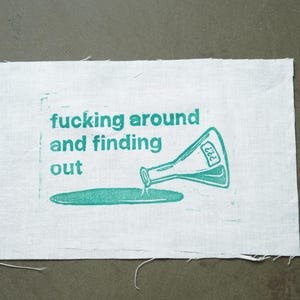 Peut inclure: Écusson en tissu blanc avec l'inscription « fucking around and finding out » en lettres turquoise. Un bécher turquoise verse du liquide sur une flaque turquoise. Bords bruts.