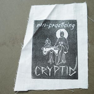 May include: A white fabric patch with a black and white graphic print. The print features a figure with a halo, seated at a table, with the words "non-practicing CRYPTID". The patch has raw edges.
