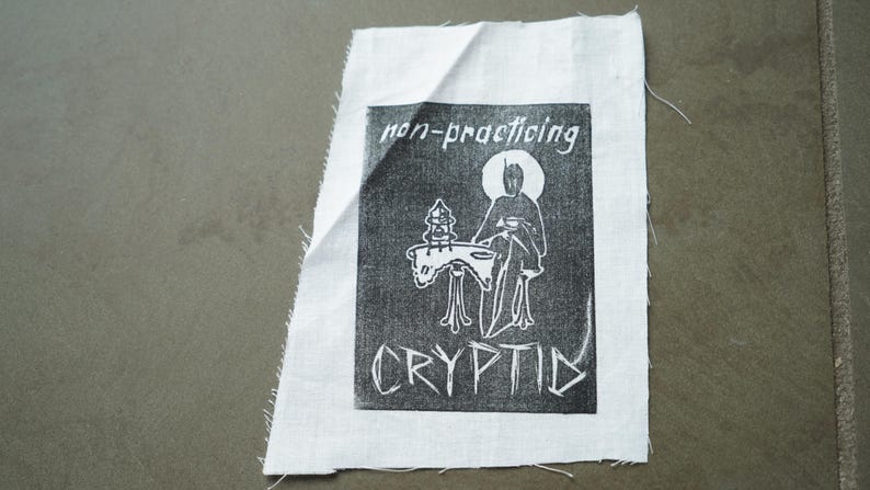 Może przedstawiać: Biała naszywka z tkaniny z czarno-białym nadrukiem graficznym. Nadruk przedstawia postać z aureolą, siedzącą przy stole, ze słowami "non-practicing CRYPTID". Naszywka ma surowe krawędzie.