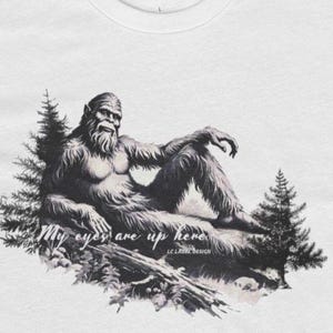 May include: A white t-shirt with a black and white illustration of a Bigfoot sitting on a log. The text "My eyes are up here" is written below the image.