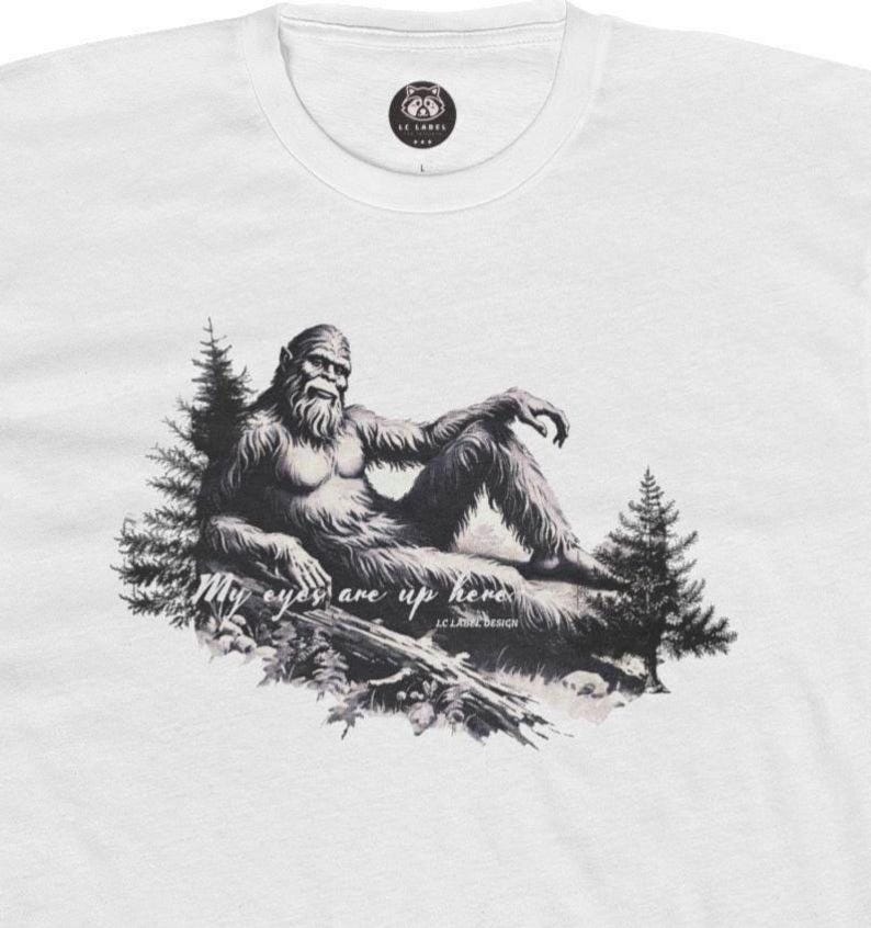 May include: A white t-shirt with a black and white illustration of a Bigfoot sitting on a log. The text "My eyes are up here" is written below the image.